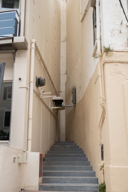 A narrow outdoor stairway with grey concrete steps ascending between two tall, beige building walls. On the left side, a small balcony with blue paneling is visible, and an electrical meter and severa