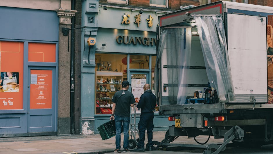 A white flatbed truck performing furniture transport and home relocation in a busy urban street with narrow roads and multiple shops on either side. The truck is loaded with wooden pallets and wrapped